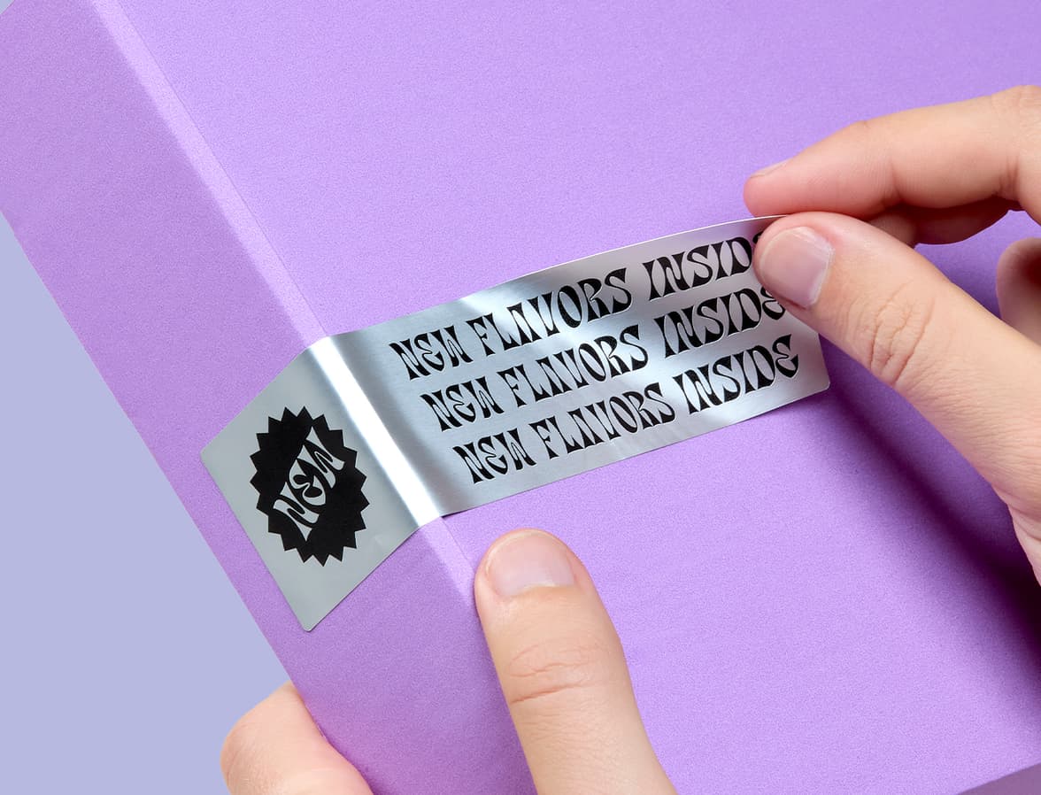 A hand placing a customized silver metallic sticker on the edge of a packaging box.