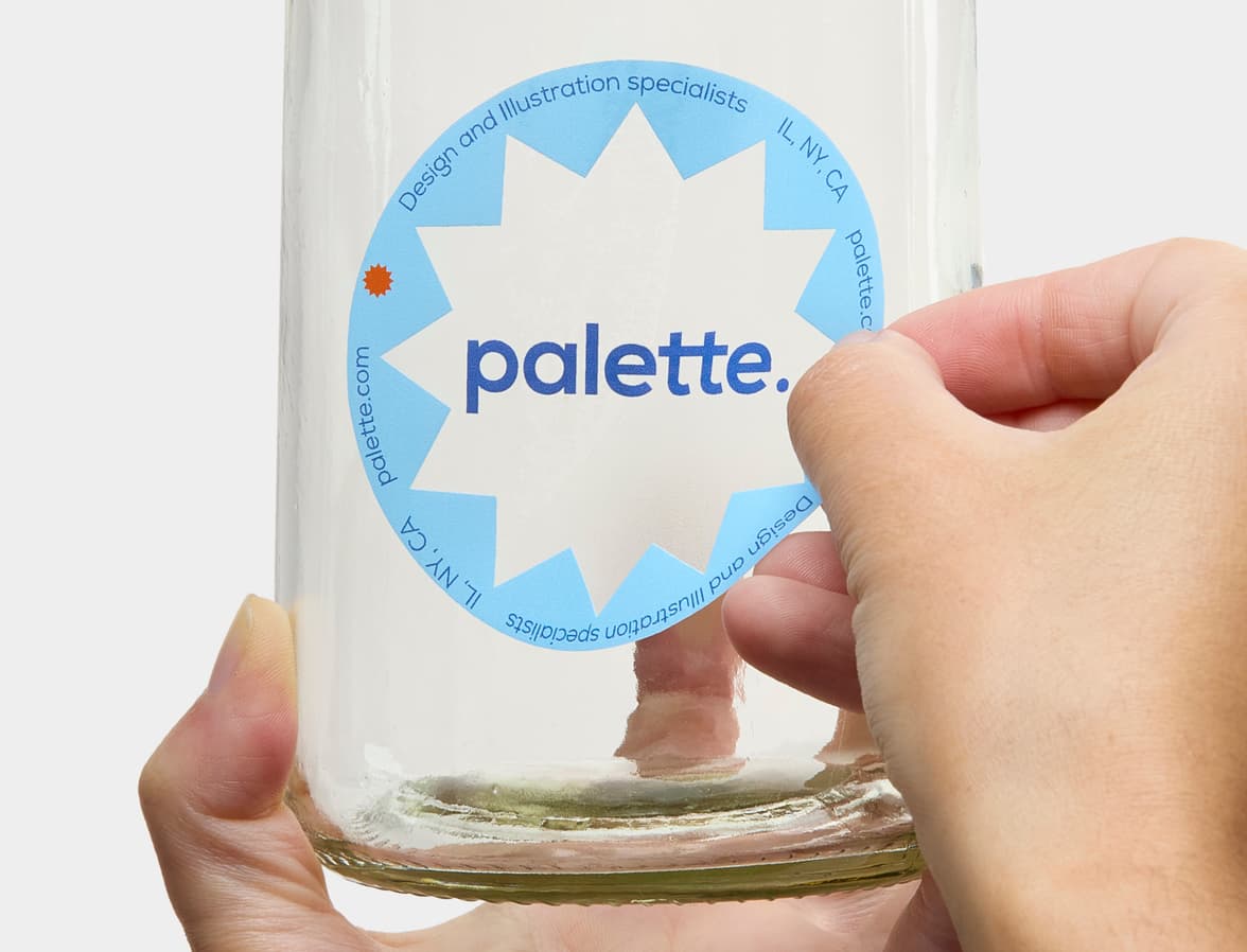 A hand placing a customized clear round sticker onto a glass bottle.