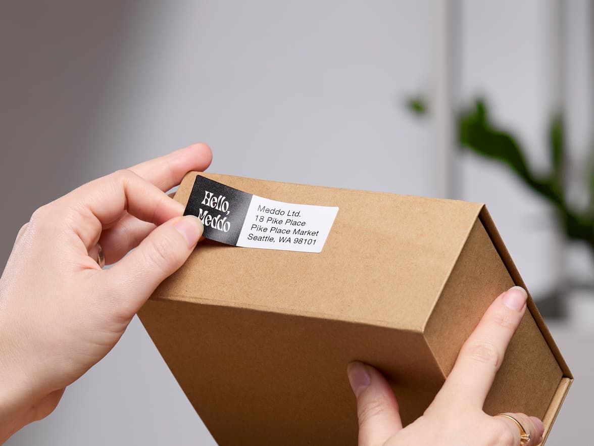 A rectangular white and black return address label being placed onto a cardboard box.