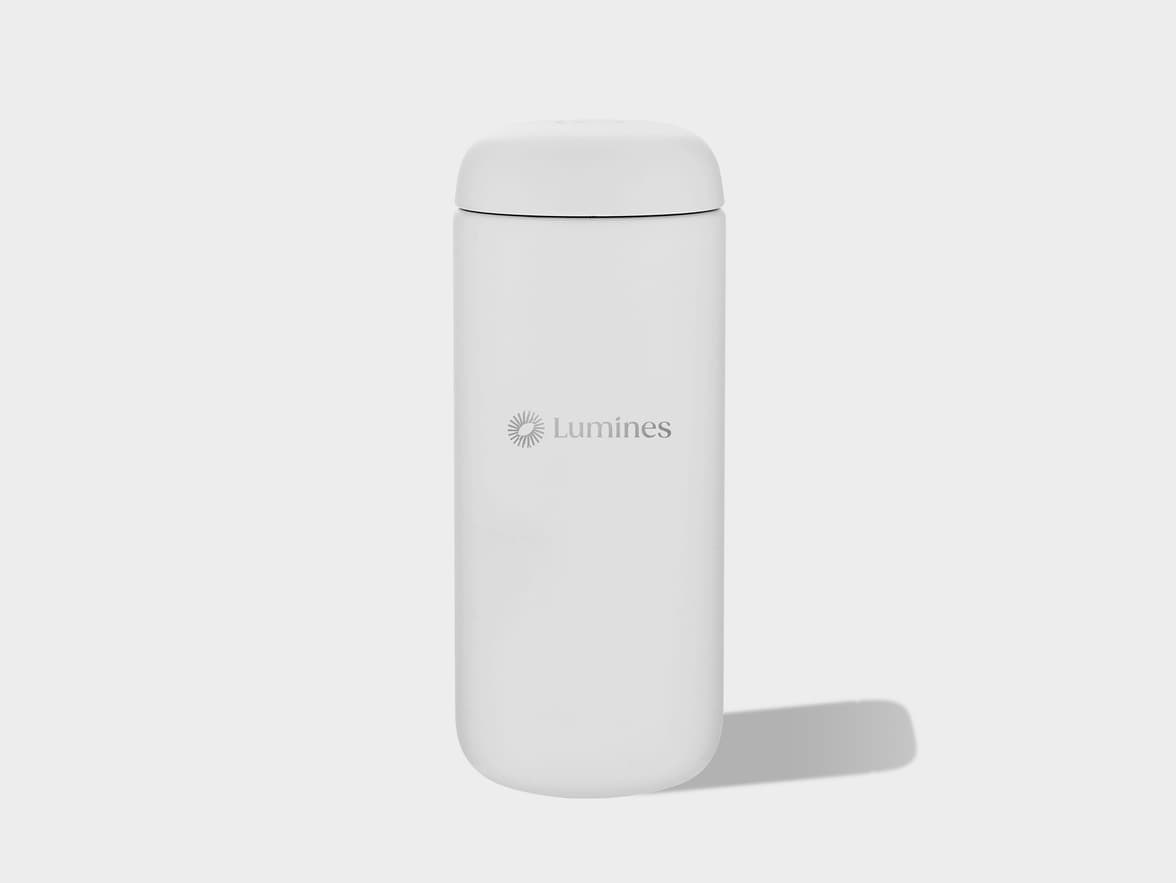 A white cylindrical insulated bottle with a rounded cap stands upright on a light gray surface, casting a shadow to its right. The "Lumines" logo, featuring a sun-like emblem and text, is visible on the front of the bottle.