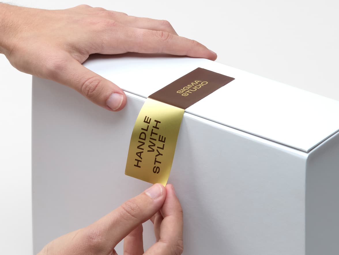 A hand placing a customized gold and brown metallic sticker on the corner of a white box.