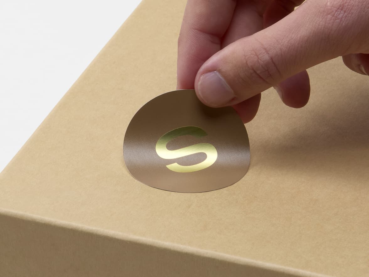A hand placing a round gold metallic sticker on a cardboard box.