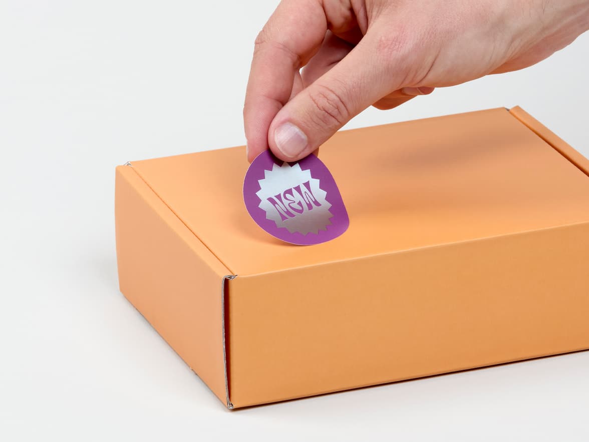 A hand placing a round silver sticker with purple customization on a orange box.