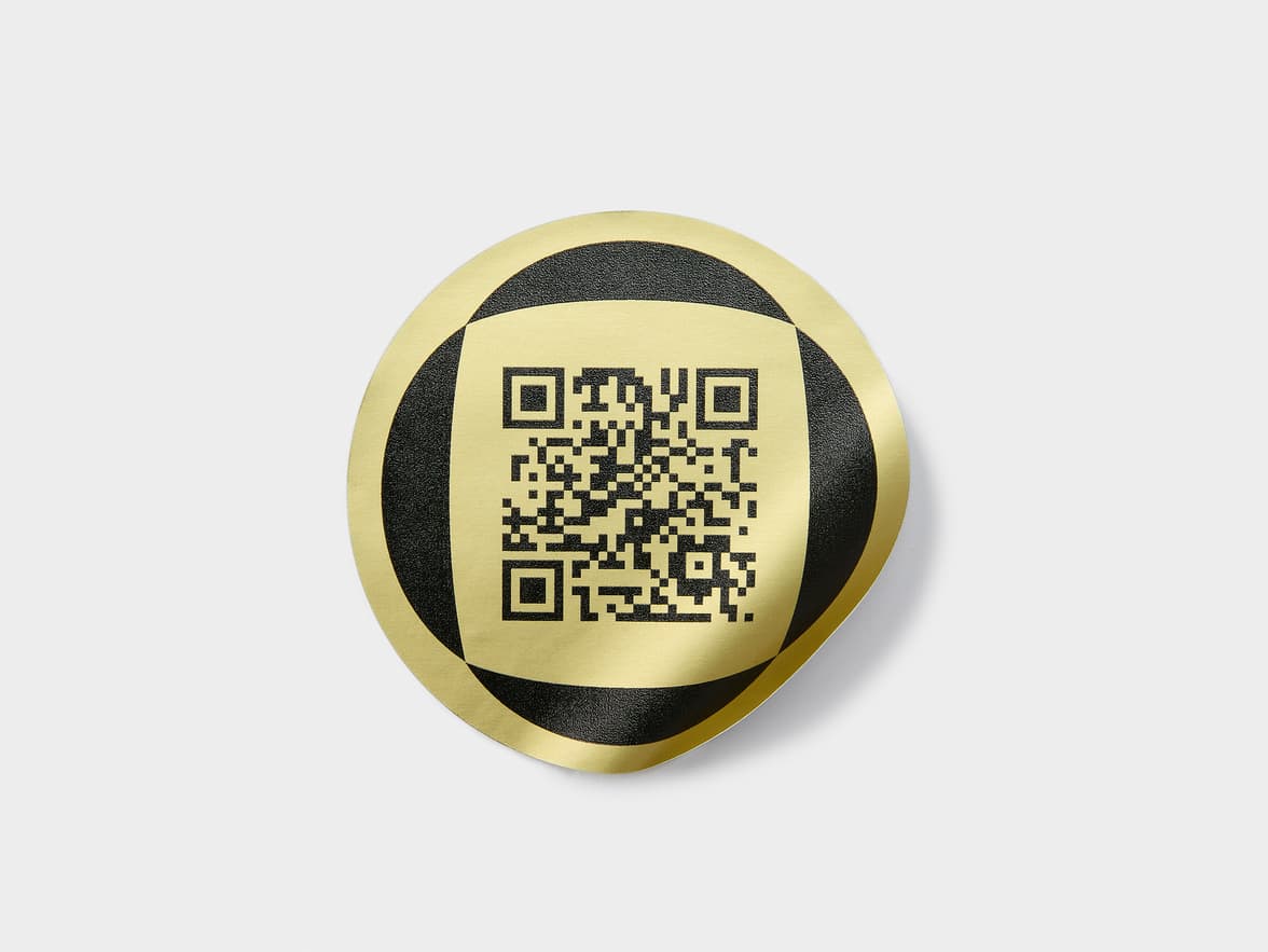 A round gold metallic foil sticker with a black QR code on it.