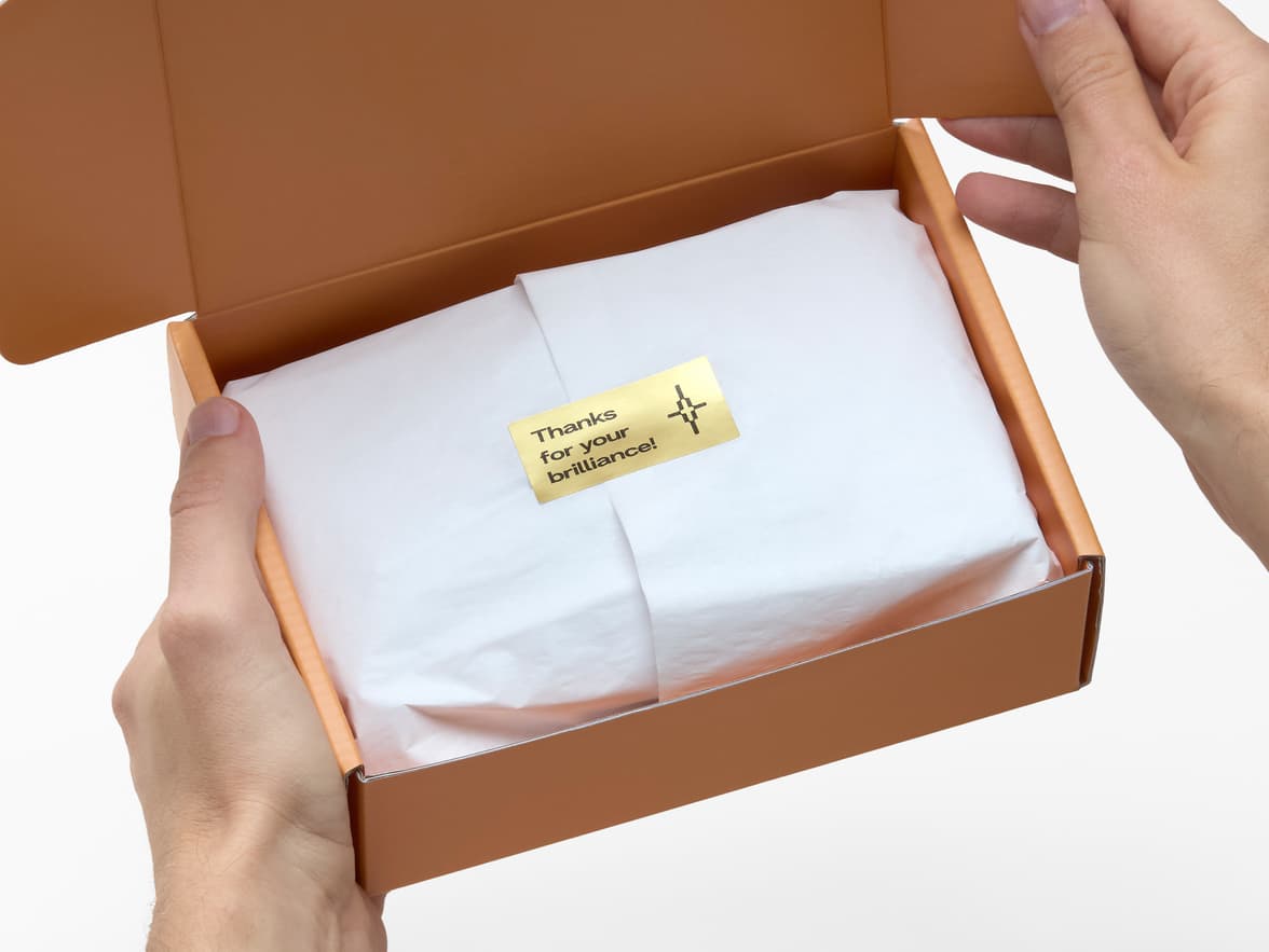 Hands holding a packaging box with white tissues inside sealed by a gold metallic sticker.