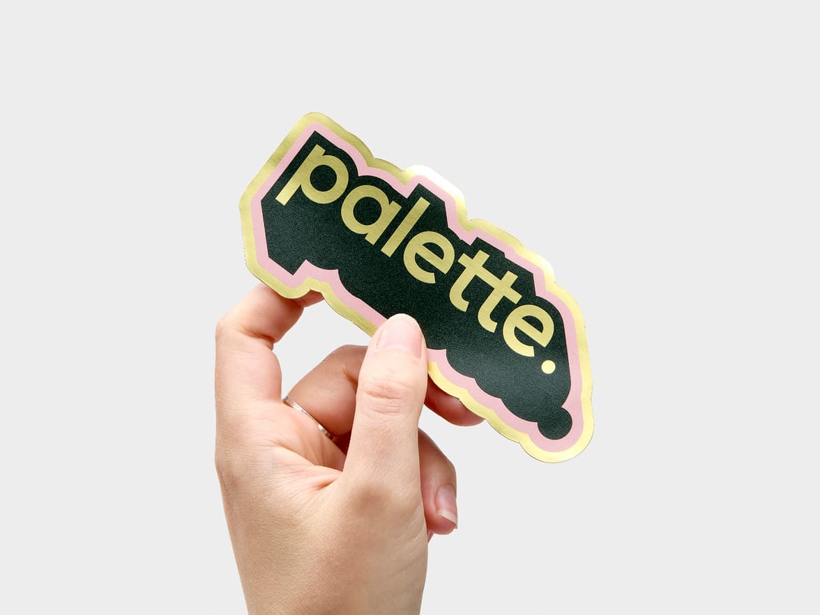 A hand holding a customized gold metallic sticker in the design and shape of the word 'palette'.
