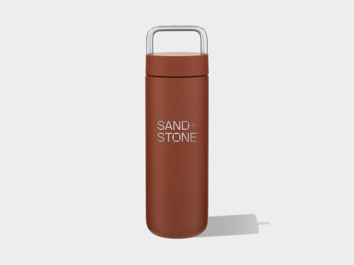 A rust-colored tumbler with a clear handle, branded "SAND + STONE".