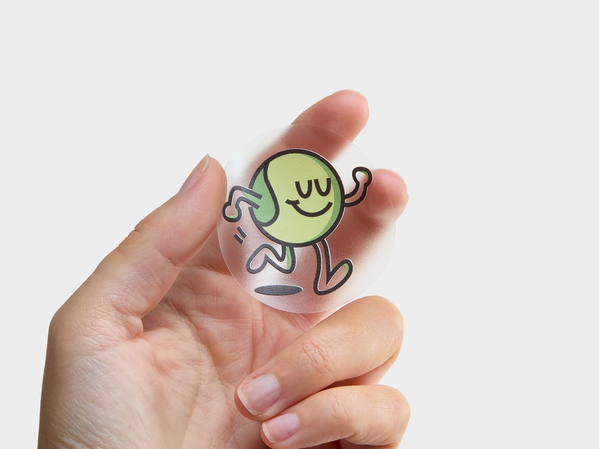 A hand holding a customized round clear sticker with a green cartoon character printed on it.