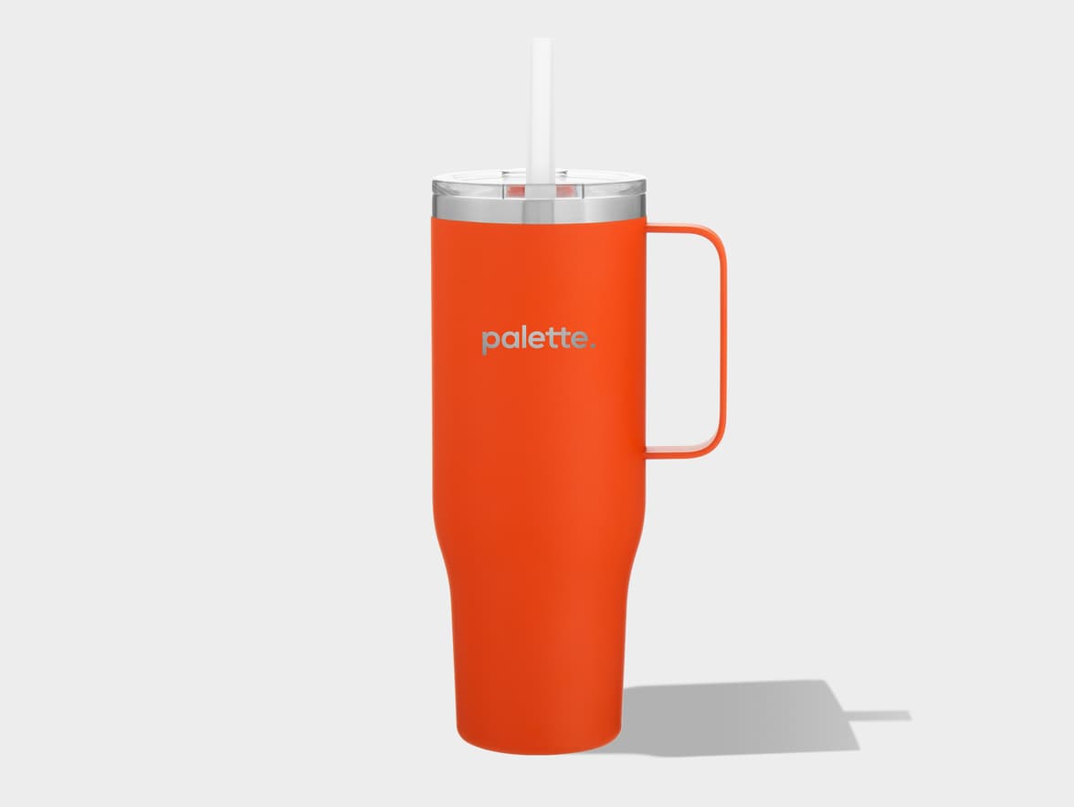 An orange travel tumbler with a handle is featured against a plain light gray background. The tumbler has a clear lid with a silver-colored rim and a white straw protruding from the top. The word "palette." is subtly embossed in a lighter gray shade on the front of the orange body. A soft shadow extends to the right from the base of the tumbler.