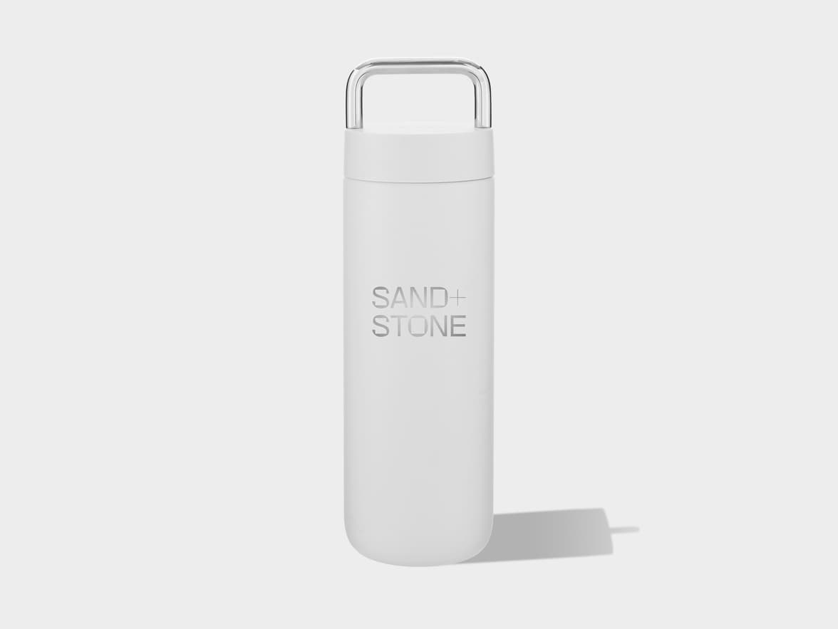 A minimalist white water bottle with a metallic U-shaped handle on top and the words "SAND+" and "STONE" in silver text on its front, casting a soft shadow on a light gray background.