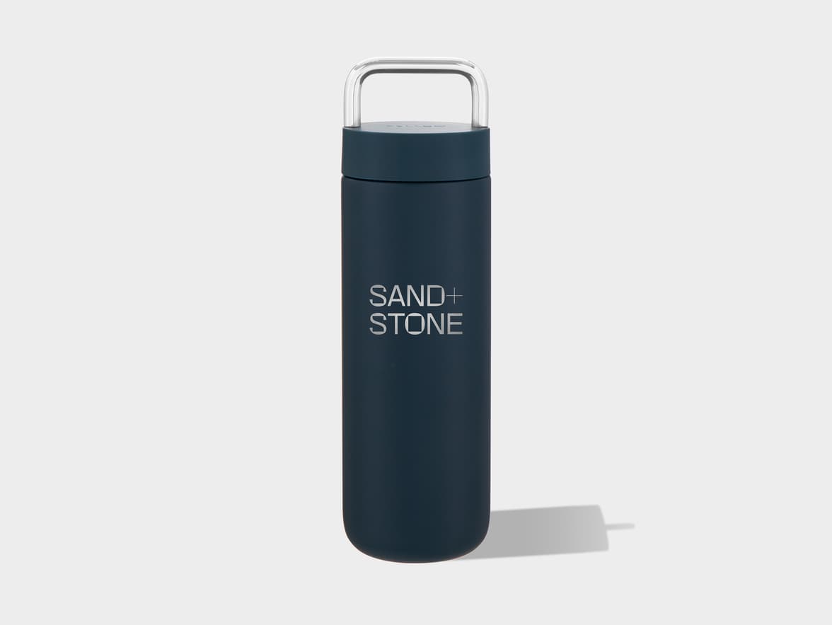 A dark blue cylindrical water bottle with a rounded base and a silver U-shaped handle on its lid, standing on a light grey surface with a shadow to its right. The words "SAND+STONE" are printed vertically in light grey on its front.