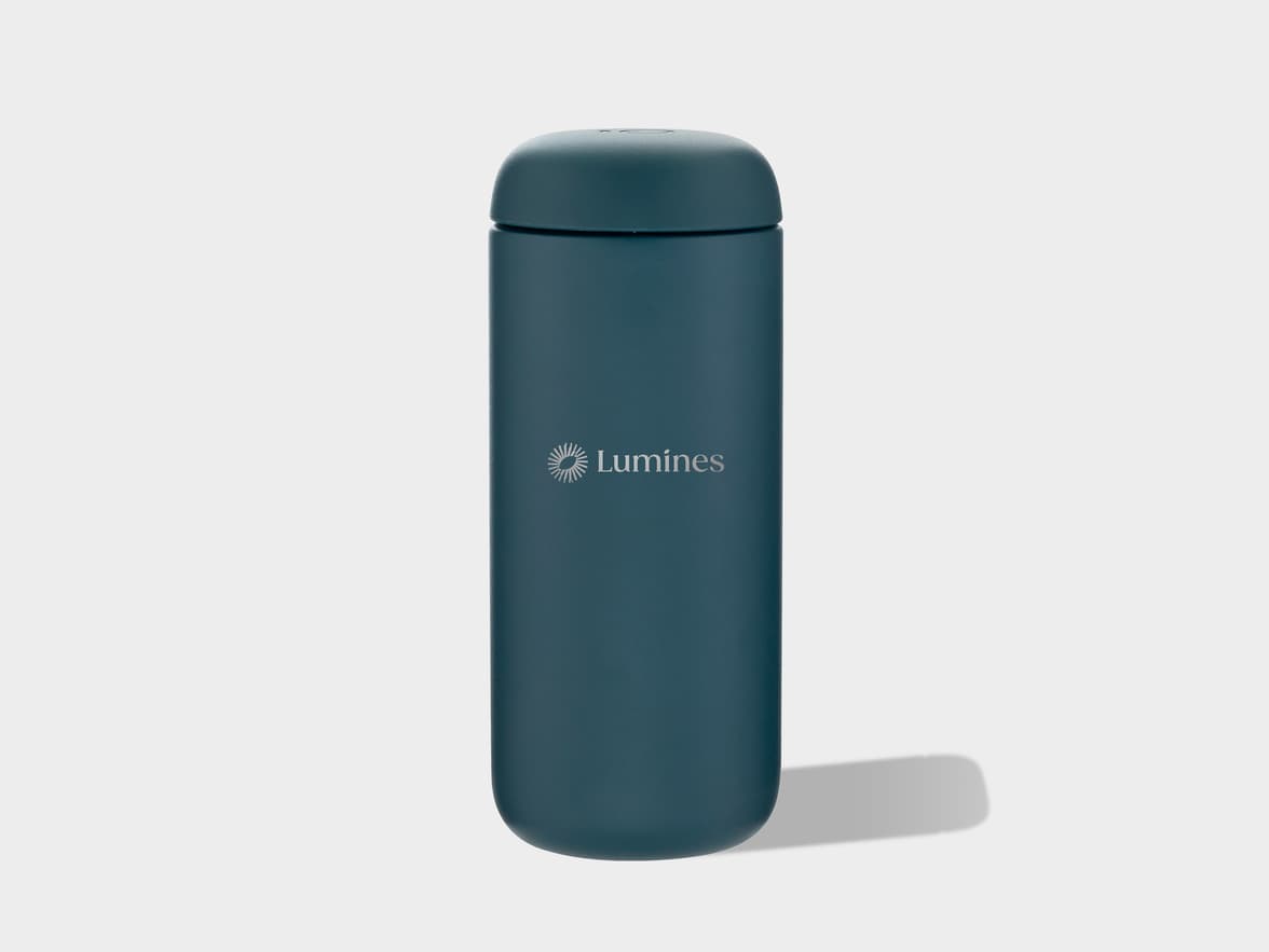 A dark blue-green (petrol blue) matte insulated tumbler with rounded top and bottom stands on a light gray surface, casting a soft shadow to its right. The front of the tumbler features a silver 'Lumines' logo with a stylized sun or starburst icon.