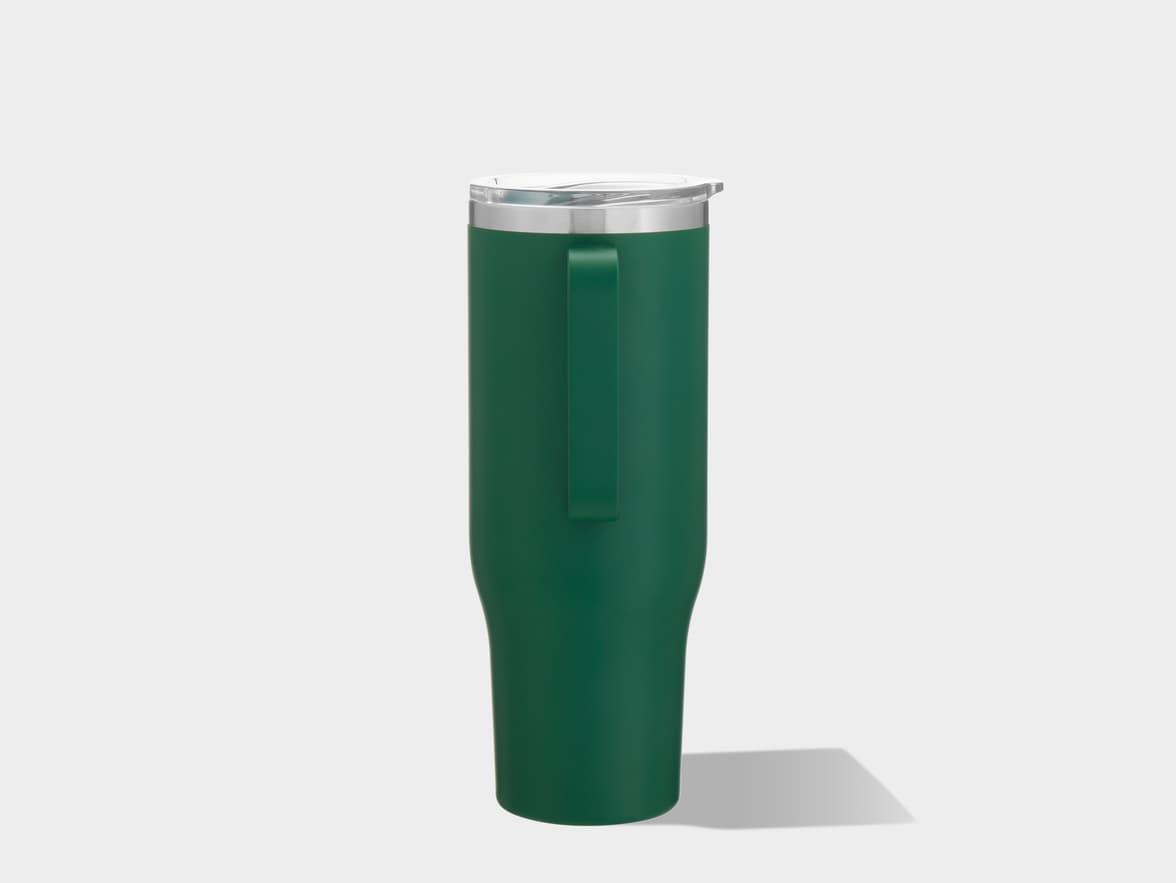 A tall, dark green matte-finished insulated tumbler with a handle is positioned upright and slightly angled to the right against a plain light gray background. The tumbler has a wider base that tapers upwards slightly before widening again near the top. A dark green, flat, rectangular handle runs vertically along one side. The top of the tumbler features a silver-colored rim and a clear, translucent plastic lid with a visible opening and a raised lip. A soft, gray shadow extends from the base of the tumbler to the right.