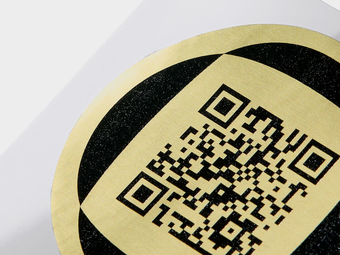 A close up image of a round gold metallic sticker with a black QR code on it.